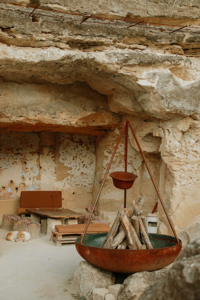 A rustic kitchen setup within a natural cave featuring a large metal fire pit.